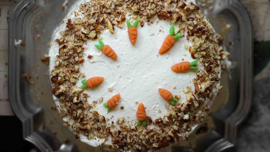 A round vegan carrot cake topped with white frosting, adorned with small fondant carrots in a circle on top. Chopped nuts line the edges, adding texture and contrast to the smooth icing.